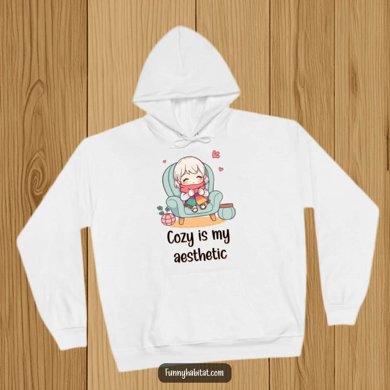 Funny hoodie depicting a cute character knitting a colorful scarf in a cozy armchair, offering warmth and humorous style for knitters.